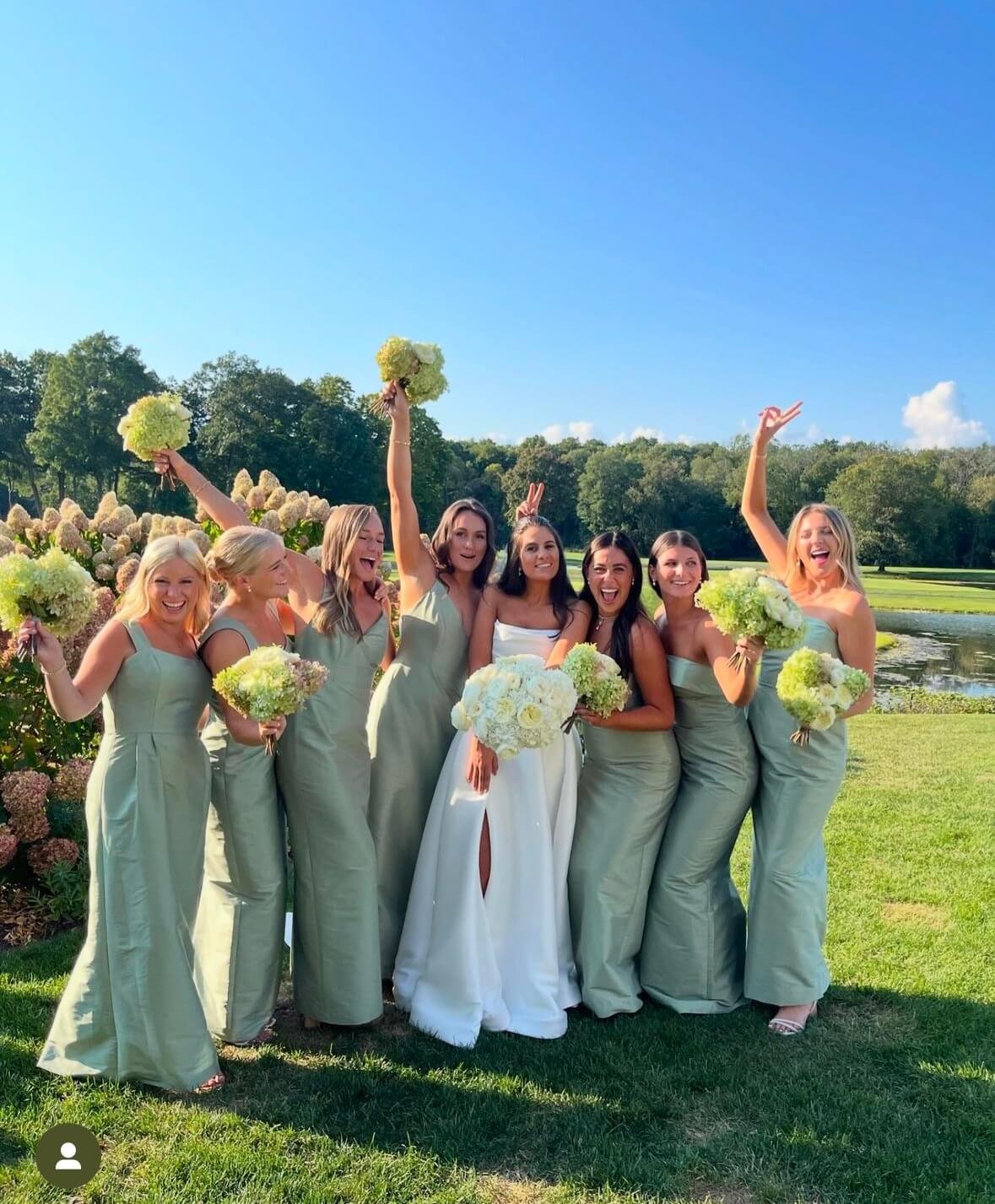 Greenwich CT makeup on a group of women in light green bridesmaid dresses and a bride in a white wedding dress holding bouquets, standing outdoors on grass with a pond and trees in the background, celebrating at a wedding