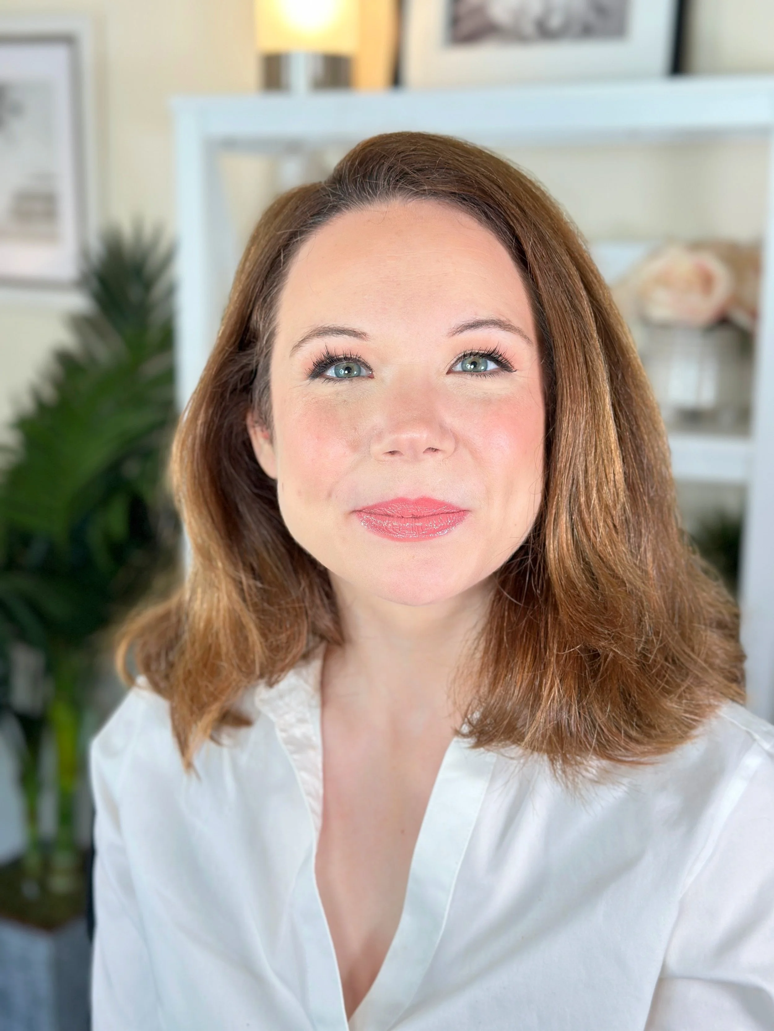 A woman with shoulder-length brown hair, blue eyes, and fair skin, wearing a white blouse, smiling slightly, indoors with plants and decor in the background.