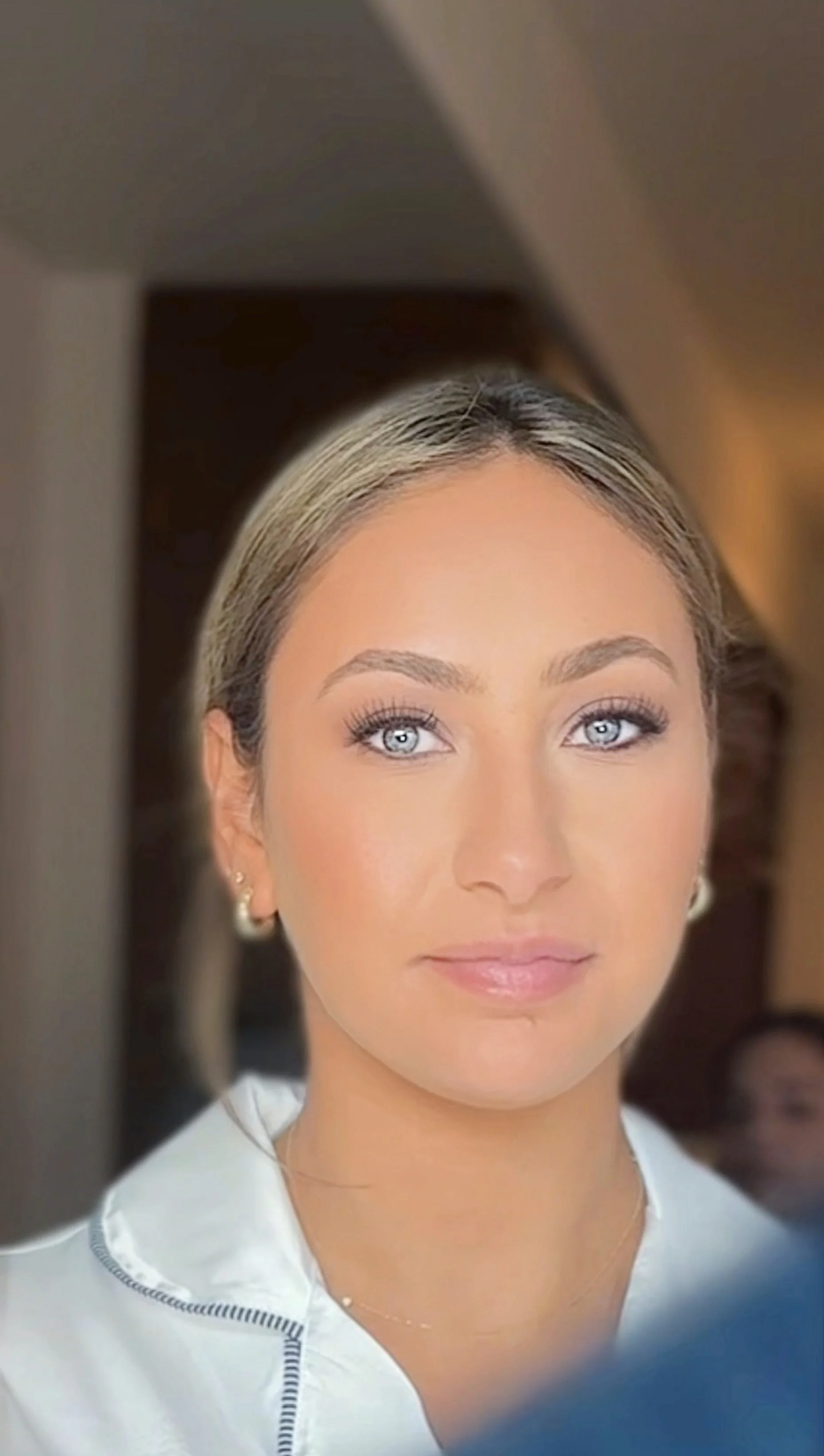 Bridal Makeup in CT with a close-up of a bridesmaid with blonde hair and blue eyes, wearing pearl earrings and a white top, smiling softly indoors.