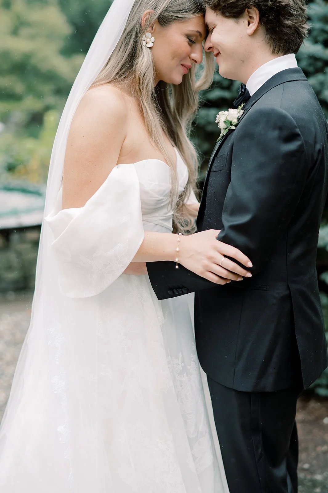 A bride and groom on their wedding day, standing close, touching foreheads, smiling with eyes closed.