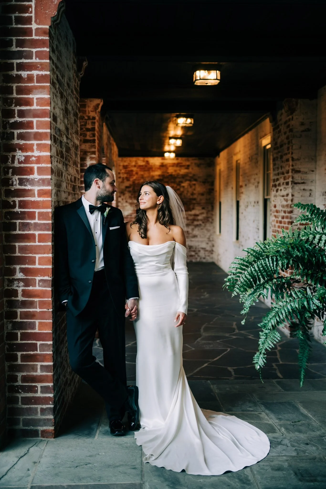 Bride with beautiful makeup in CT and groom at New Haven Lawn club sharing a moment, standing in a brick corridor with warm lighting, the bride in an off-shoulder white wedding gown, and the groom in a black tuxedo, holding hands.
