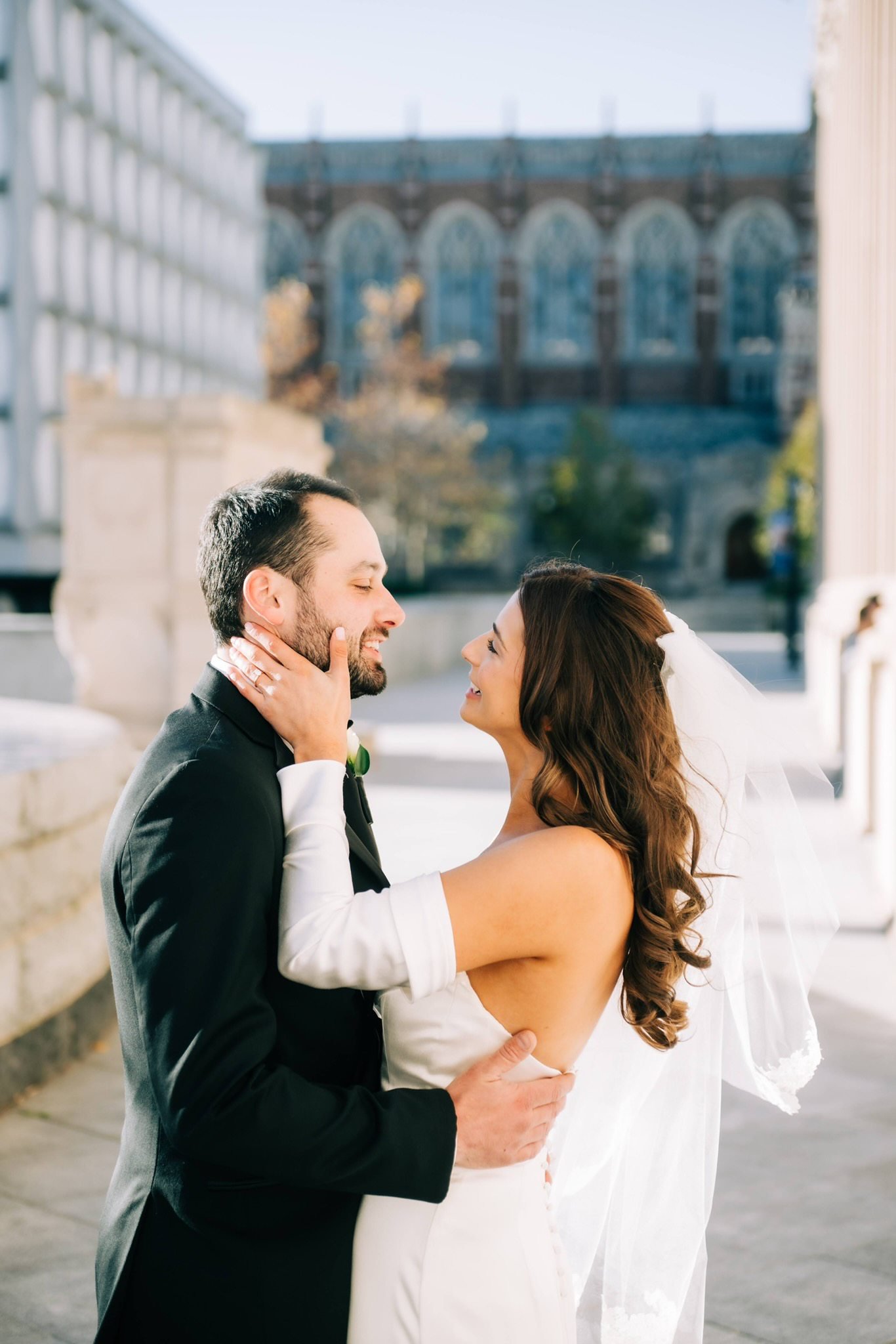 A bride and groom in wedding attire sharing a moment outdoors, with the bride holding the groom's face and the groom holding the bride's waist, smiling at each other.
