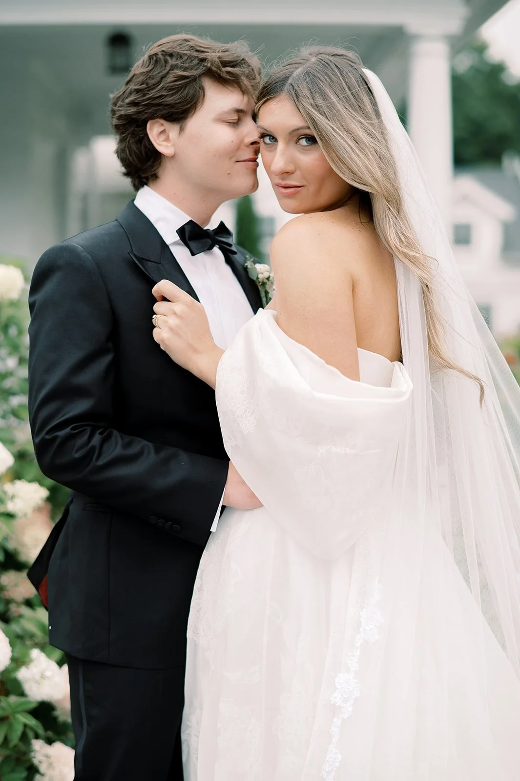 A bride with beautiful wedding makeup and groom in Newtown, CT in wedding attire sharing an intimate moment outdoors, with the groom whispering into the bride's ear and the bride looking at the camera, surrounded by greenery and flowers.