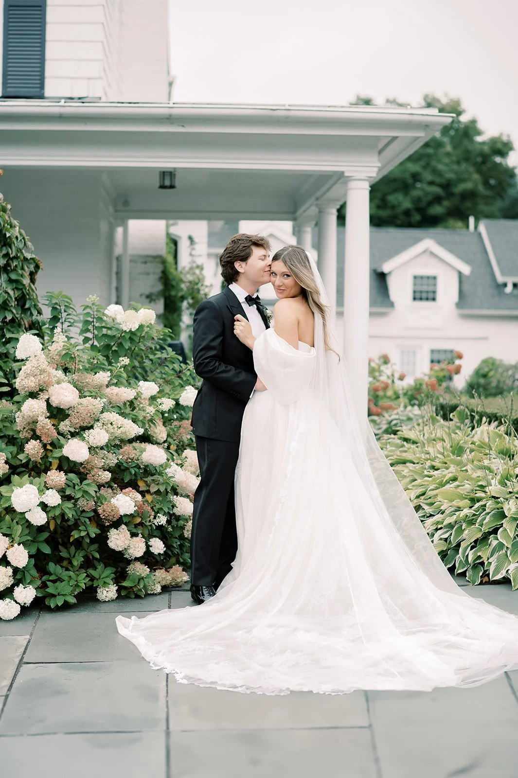 A CT bride with beautiful makeup and groom standing together outdoors in front of a white house with columns, surrounded by flowers, with the bride wearing a white wedding dress and the groom in a black tuxedo, posing for a wedding photo.