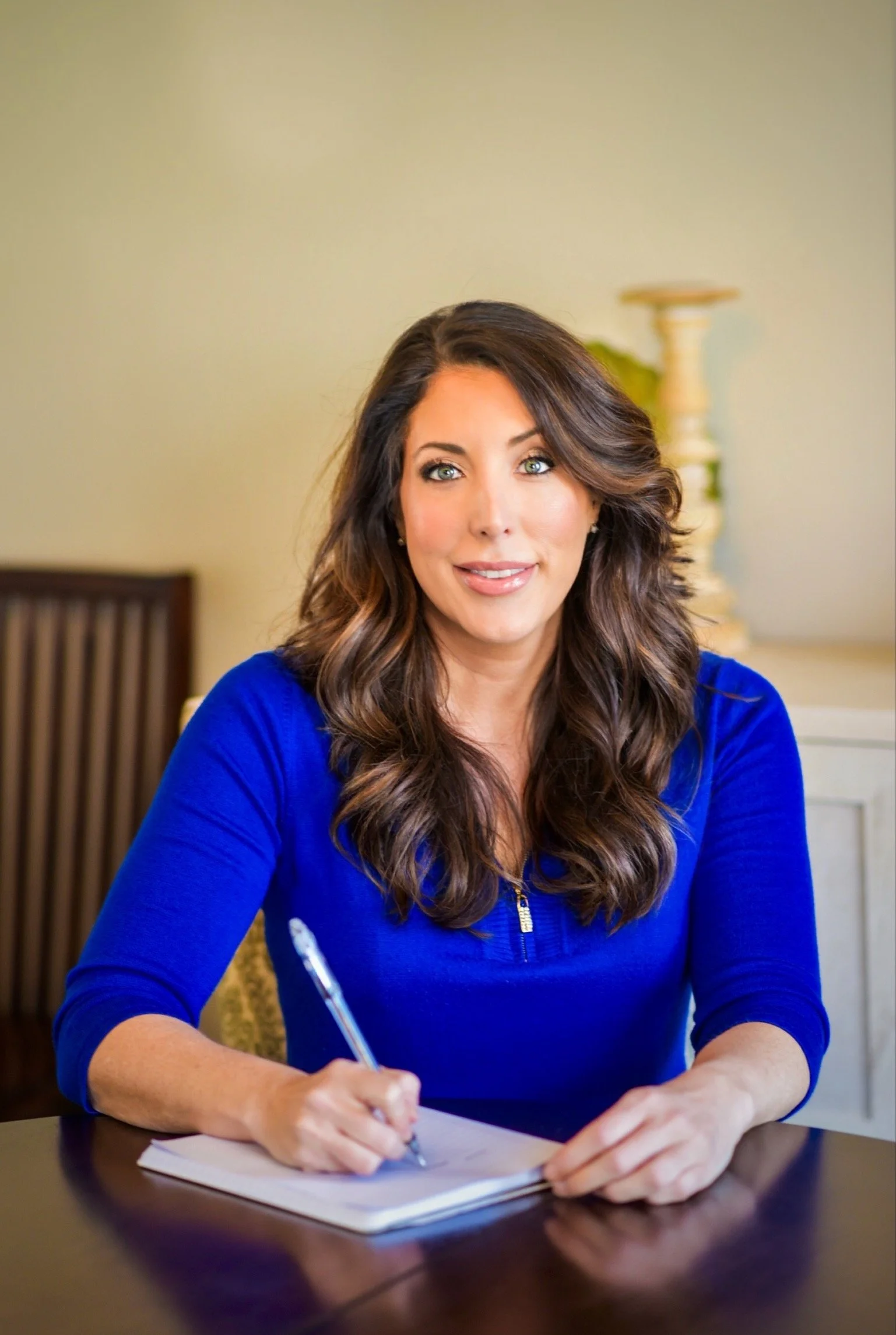 Makeup Artist Fairfield CT woman with long wavy brown hair and blue eyes, wearing a royal blue top, sitting at a table, holding a pen, smiling, in a warmly lit room.