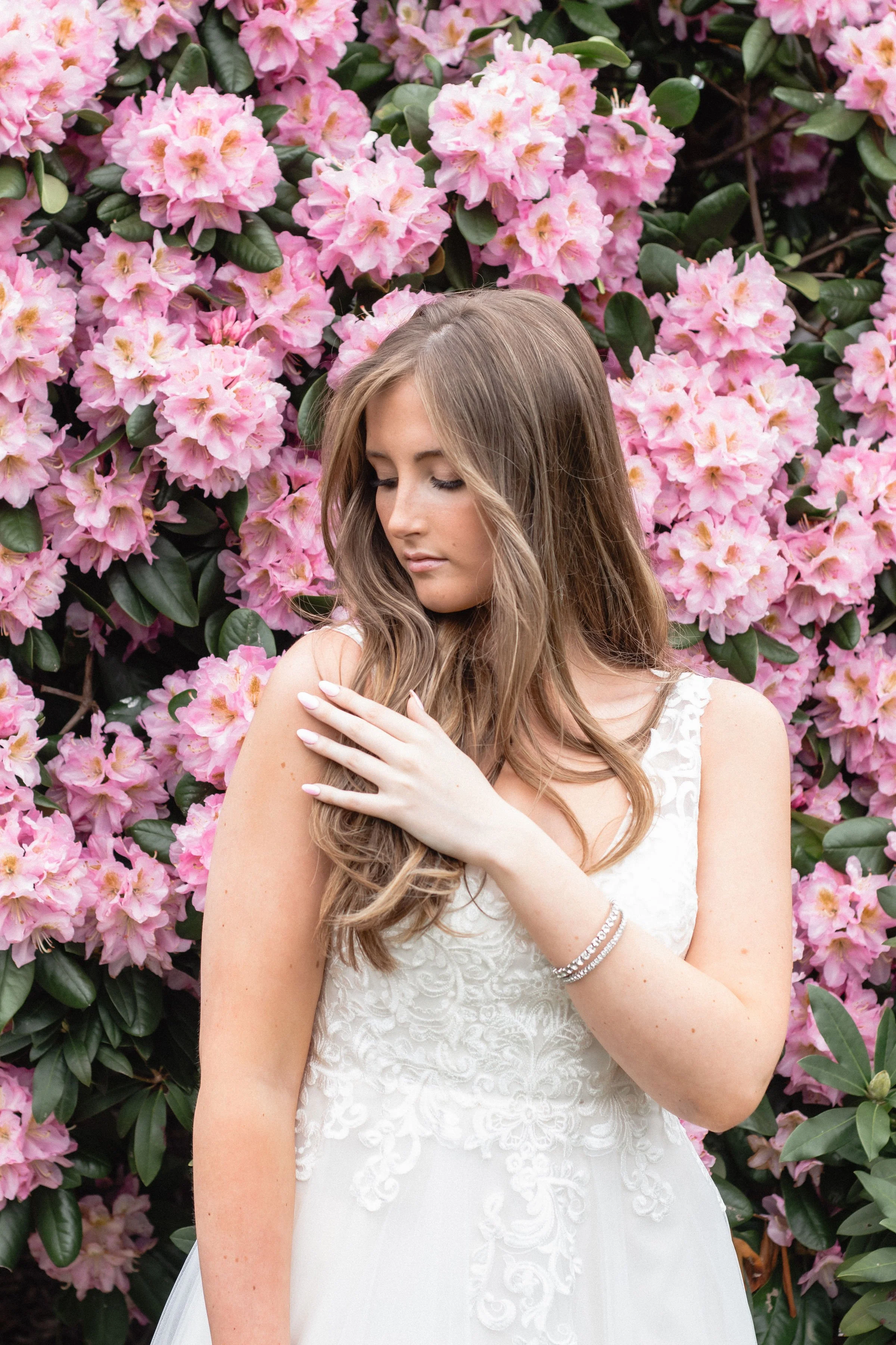 A young woman in a white lace dress standing in front of pink rhododendron flowers, with her eyes closed and one hand gently resting on her shoulder.