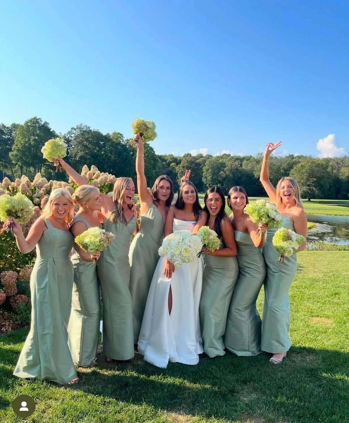 CT Wedding Makeup at Burning Tree Country Club in Greenwich CT with green dresses and a bride in a white dress holding bouquets, celebrating outdoors on a sunny day near a pond with trees in the background.