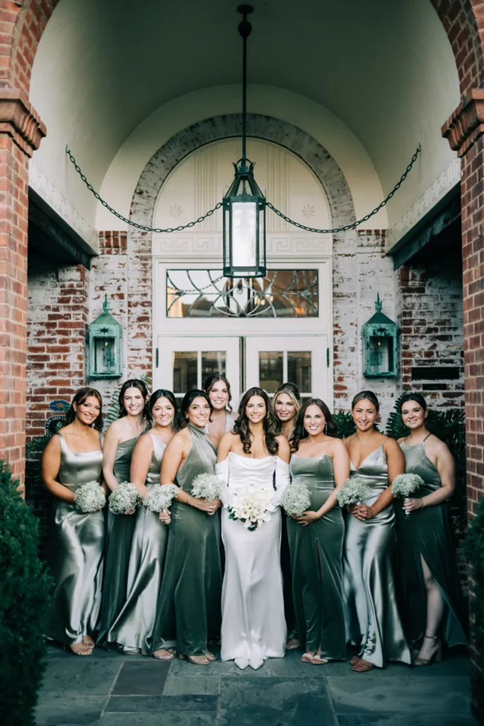 Makeup Artist Fairfield CT with wedding party of nine women in New Haven, CT including a bride in white and bridesmaids in matching silver dresses, posing together outside a brick building with a large lamp hanging overhead.