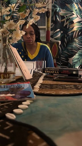 A quiet, intimate scene of a woman reading at a patterned table, surrounded by books, flowers, and cultural objects, set against lush botanical wallpaper, evoking reflection, heritage, and calm focus.