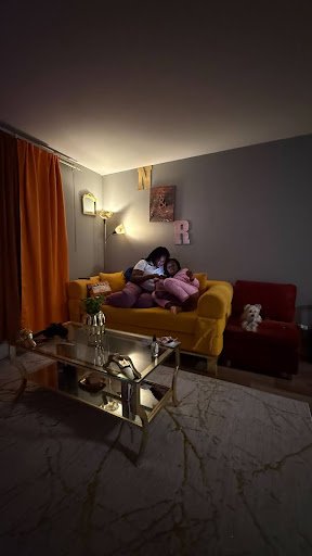 Two figures rest closely on a mustard-yellow sofa, absorbed in shared stillness, as warm lamplight and domestic details frame an intimate scene of care, comfort, and quiet connection within a lived-in home.