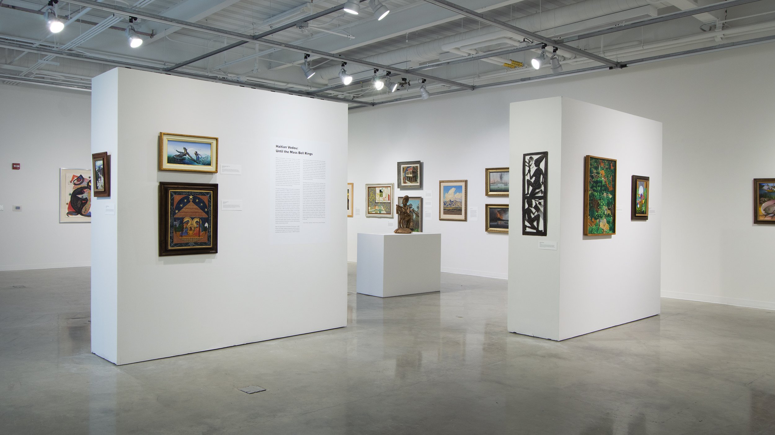 A view of an exhibition of Haitian art. Two floating walls stand adjacent, each displaying paintings of Haitian subjects and scenes. The far walls display additional paintings of American landscapes and post-War modernist prints.