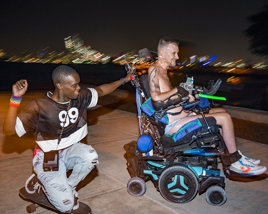    They see us rollin'   ,  2022 Digital pigment print on Slickrock Metallic Pearl 260 paper 40 in x 53 inches  A side-view of Robert, a white man with shaved blonde hair, smiling in his motorized wheelchair with a smiling Black man on a skateboard t
