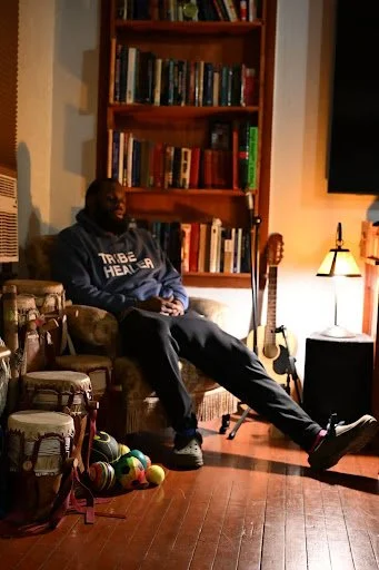 In a softly lit room of books and instruments, a resting figure is surrounded by drums, strings, and quiet rhythms, evoking reflection, healing, and lived ritual.