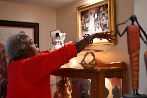 An elder reaches toward a framed family portrait, her gesture suspended among treasured objects, where memory, lineage, and care quietly fill the room.