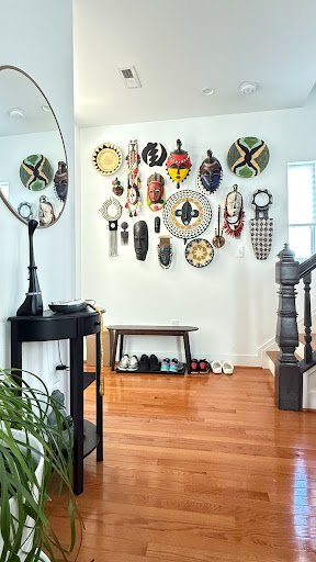 An entryway transformed into a quiet gallery: carved masks and woven forms gather on white walls, grounding the domestic space in memory, craft, and cultural presence.