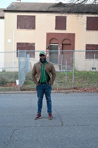 A solitary figure stands confidently in the street before a boarded, weathered building, the muted urban backdrop emphasizing themes of resilience, presence, and quiet strength.
