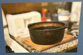 Photograph of a black, speckled ceramic bowl on a wooden cutting board atop a granite kitchen counter. Background includes a white toaster, blurred kitchen utensils, and a red object.