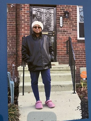 A figure stands at the threshold of a brick home, relaxed and self-possessed, where everyday attire and posture quietly affirm dignity, resilience, and belonging.