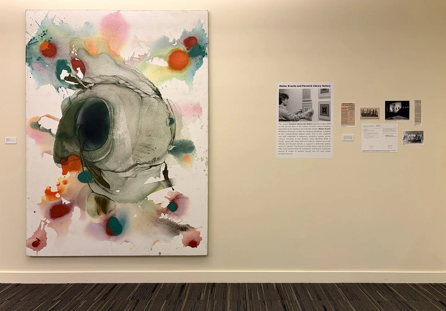 A large abstract painting in thin washes of green, purple, and orange, with deep green voids that resemble eyes and a nose. Next to the canvas are small newspaper clippings and a panel of text with a black & white photo of a man hanging a painting.