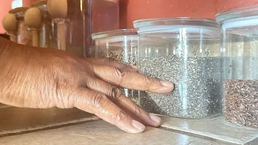 A close, tactile moment of a hand reaching toward glass jars of seeds, evoking nourishment, care, and the quiet continuity of daily ritual.