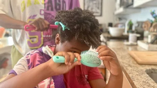A tender domestic ritual unfolds as a child concentrates on brushing textured hair, hands mid-gesture, while another figure assists just beyond focus—an intimate moment of care, patience, and shared everyday labor.