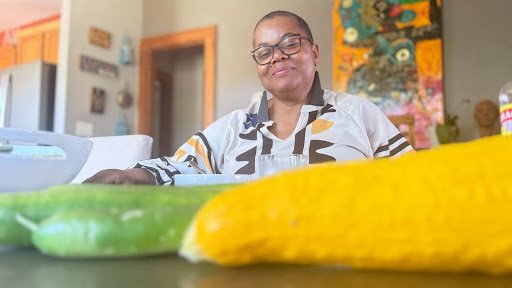 A softly lit domestic portrait captures a seated figure at the table, framed by vibrant food and patterned surroundings, conveying warmth, care, and the quiet intimacy of everyday nourishment.