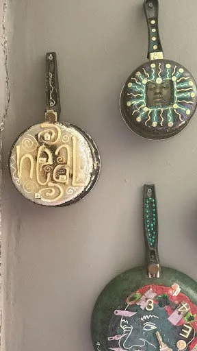 Three altered frying pans hang like icons on the wall, transforming everyday tools into symbolic portraits that merge domestic labor, spirituality, and personal affirmation.