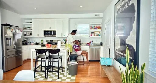 In a sunlit kitchen, a quiet moment of care unfolds as an adult braids a child’s hair, transforming an everyday domestic space into a scene of intimacy, tenderness, and inherited ritual.