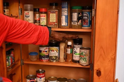 A hand hovers inside a wooden pantry, pausing among jars and cans of well-used spices—an intimate still life of nourishment, memory, and the quiet rituals of daily sustenance.