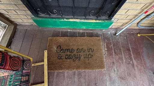 A worn welcome mat at a brick doorway bears a quiet invitation, its faded lettering and scuffed porch floor evoking warmth, humor, and the lived rhythms of home.