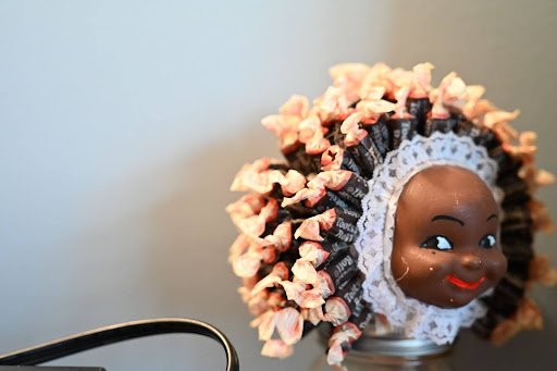 A small doll’s head adorned with a halo of candy wrappers, blending childhood nostalgia and assemblage to explore identity, adornment, and reclaimed materials.