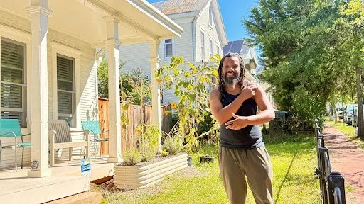 A figure stands in warm daylight before a porch and garden, where gesture, growth, and neighborhood architecture converge in a moment of ease and presence.