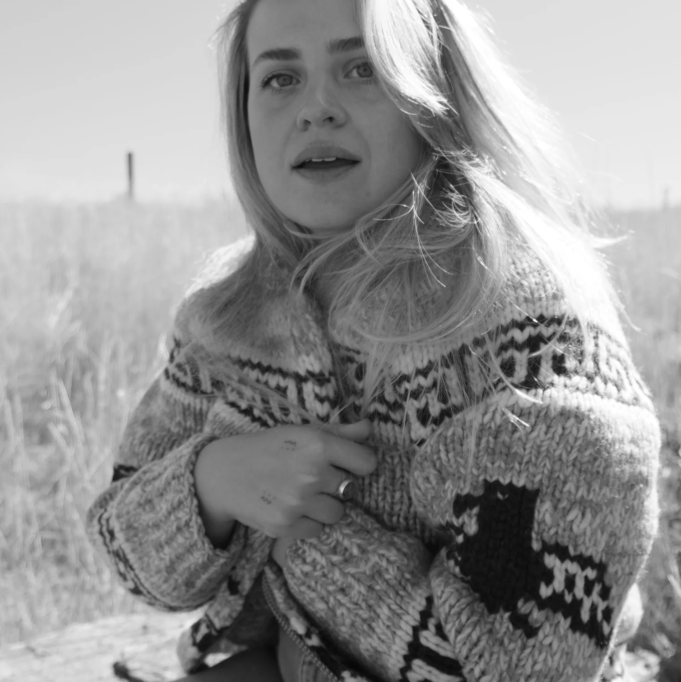 Black and white photo of a woman with blonde hair, wearing a patterned sweater, standing outdoors in a grassy area, looking at the camera with a thoughtful expression.