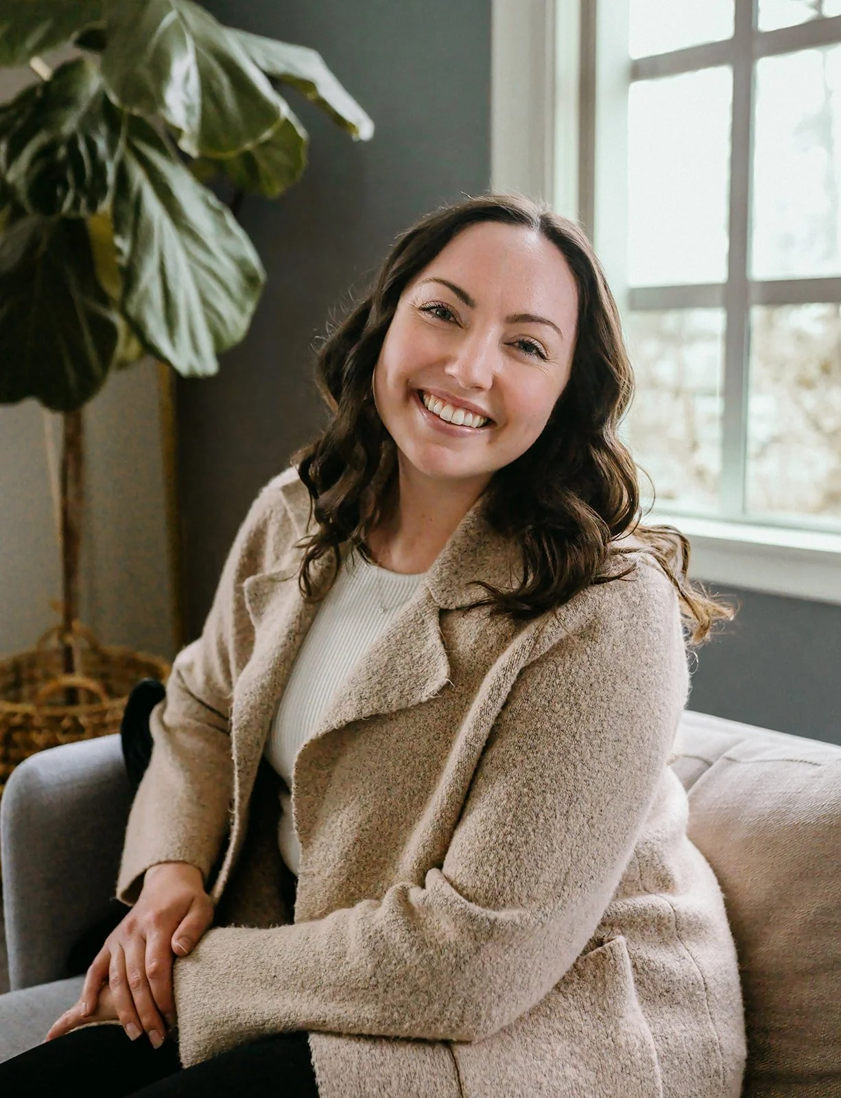 A woman with long, dark, curly hair smiling and sitting on a beige sofa in a room with natural light coming through a large window. She is wearing a light beige coat and a white top.