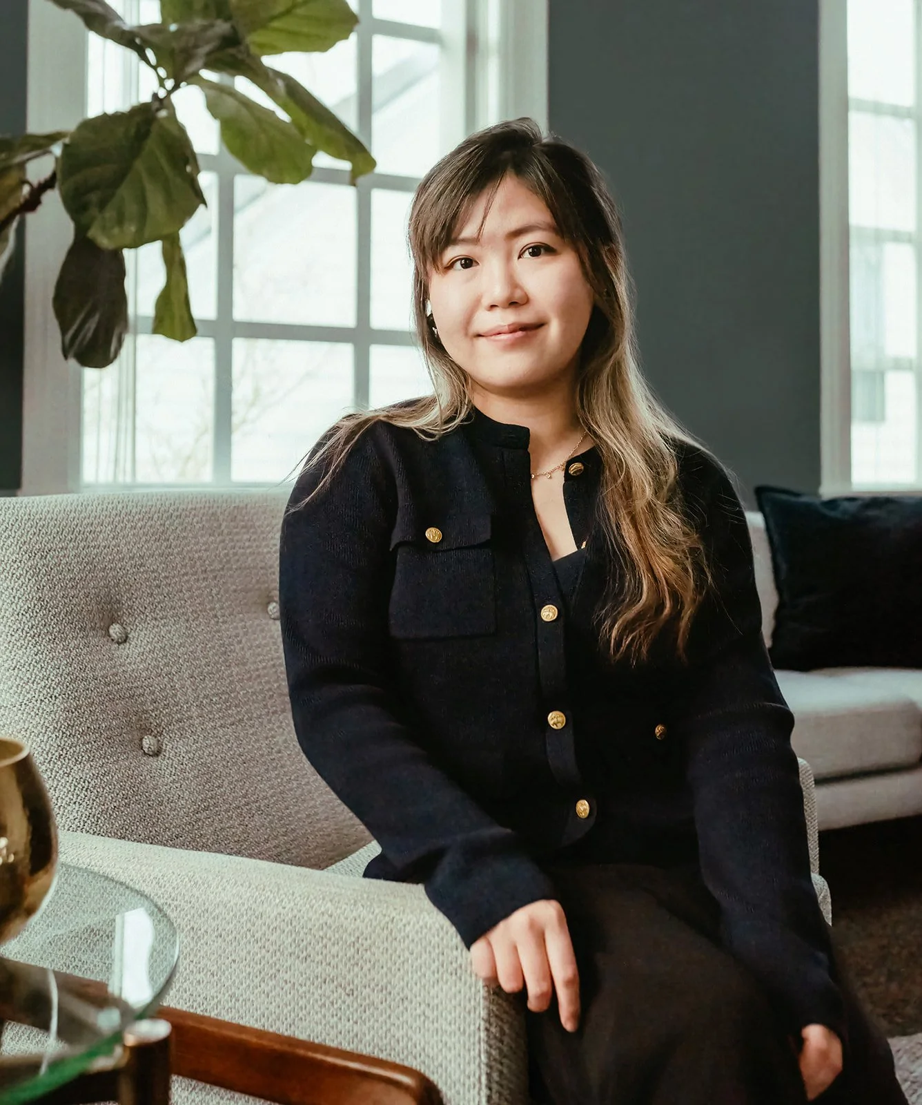 A woman with long wavy hair, wearing a black shirt with gold buttons, sitting on a beige sofa in a room with large windows, a plant in the corner, and a black cushion behind her.