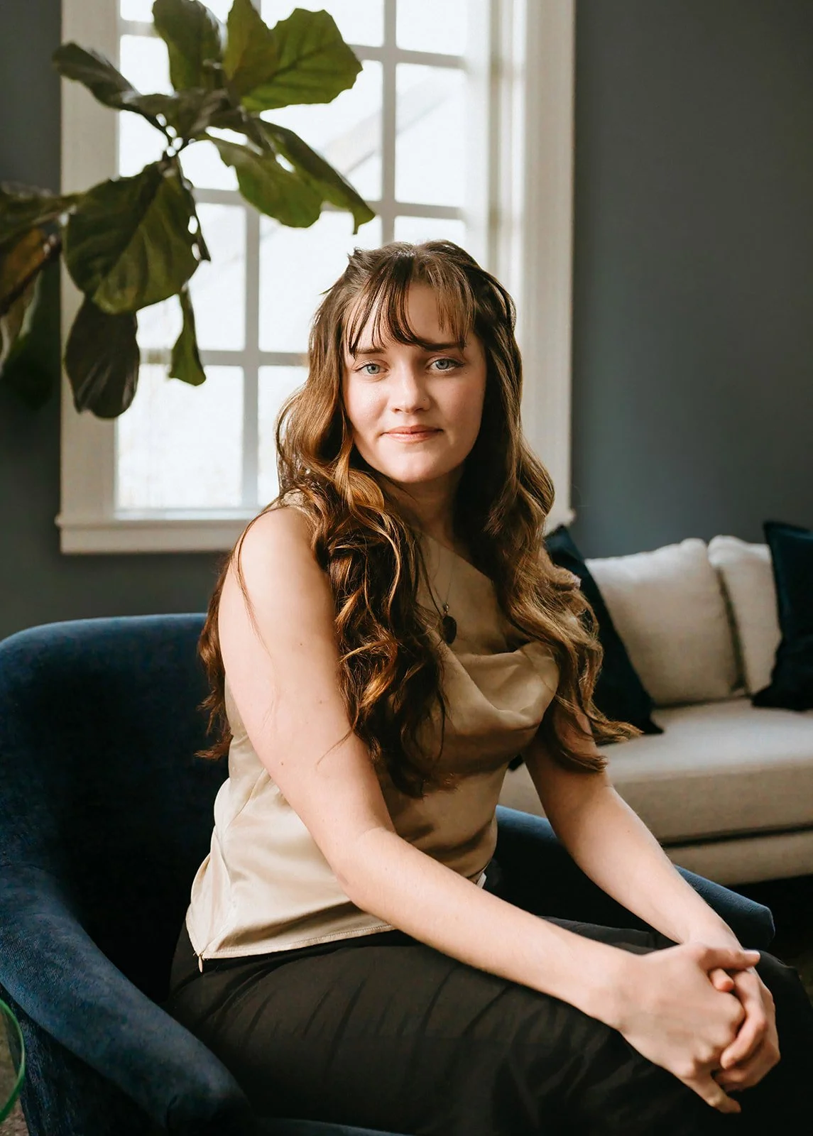 A woman with long, wavy brown hair and blue eyes sitting on a dark blue armchair in a room with large window and dark gray walls.