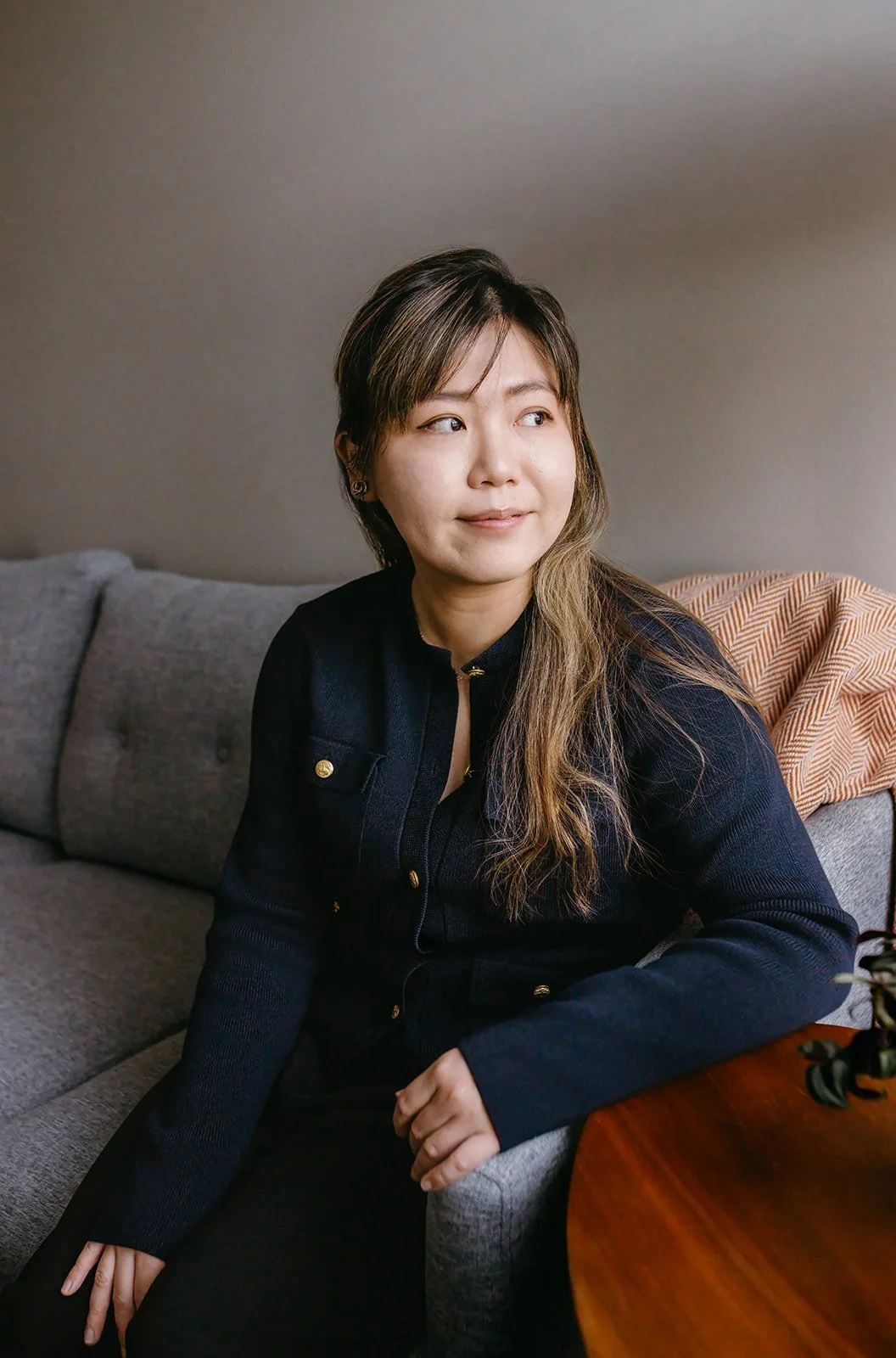 A woman with long brown hair seated on a gray sofa in front of a plain wall, looking slightly to her left, wearing a dark jacket with gold buttons.