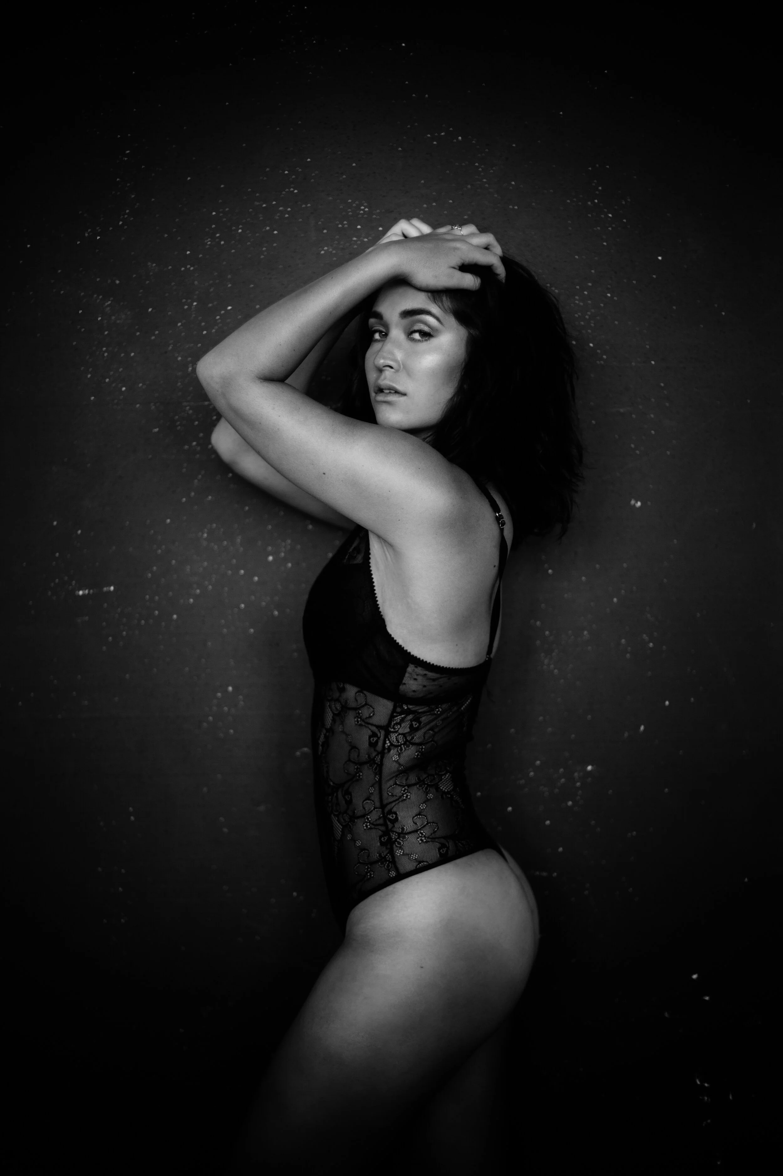 Black and white portrait of a woman posing in a lacy bodysuit against a dark background.