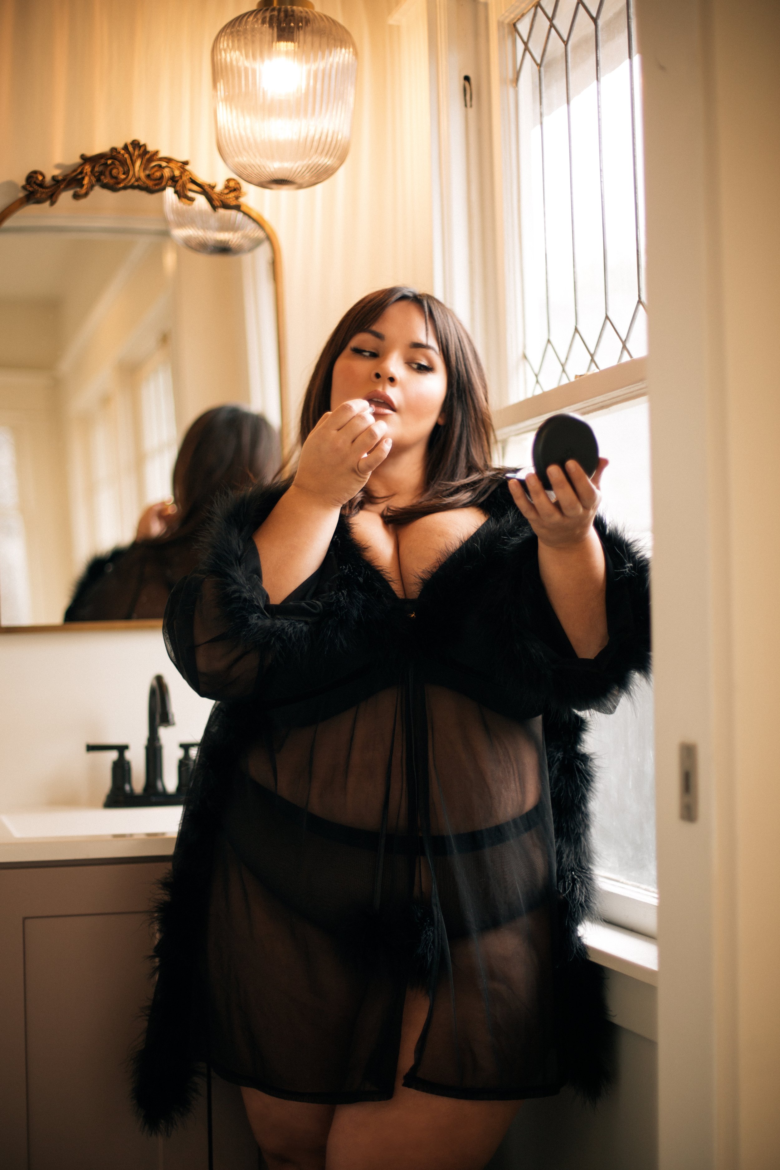 Woman applying makeup in a bathroom, wearing a sheer black robe, holding a compact mirror.