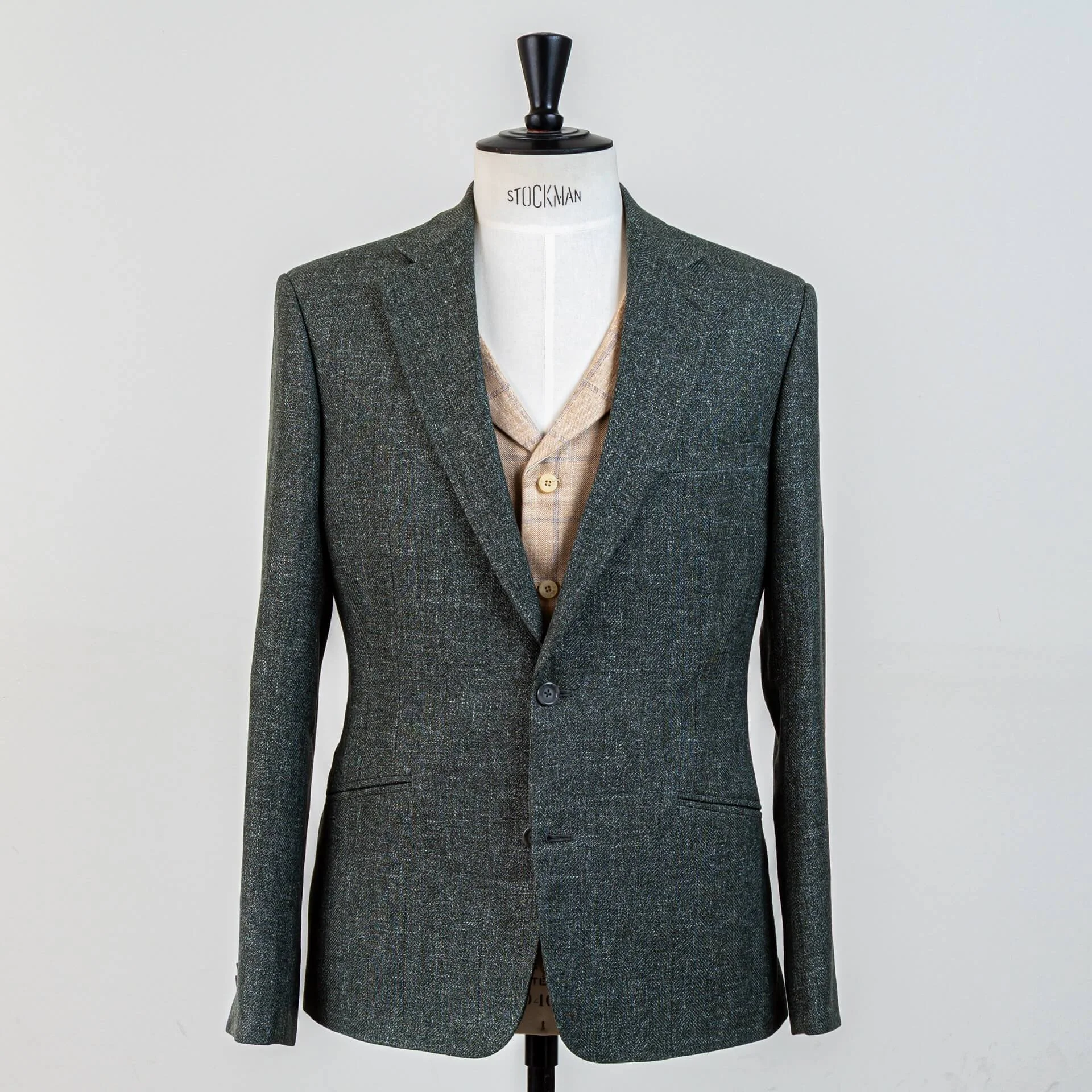 Groen 3-delig trouwpak op maat in wol-linnen-zijde blend van Piacenza, gecombineerd met zandkleurig vest met blauw ruitpatroon. Complete outfit voor zomerbruiloft, getoond op Stockman mannequin bij De Oost Amsterdam.