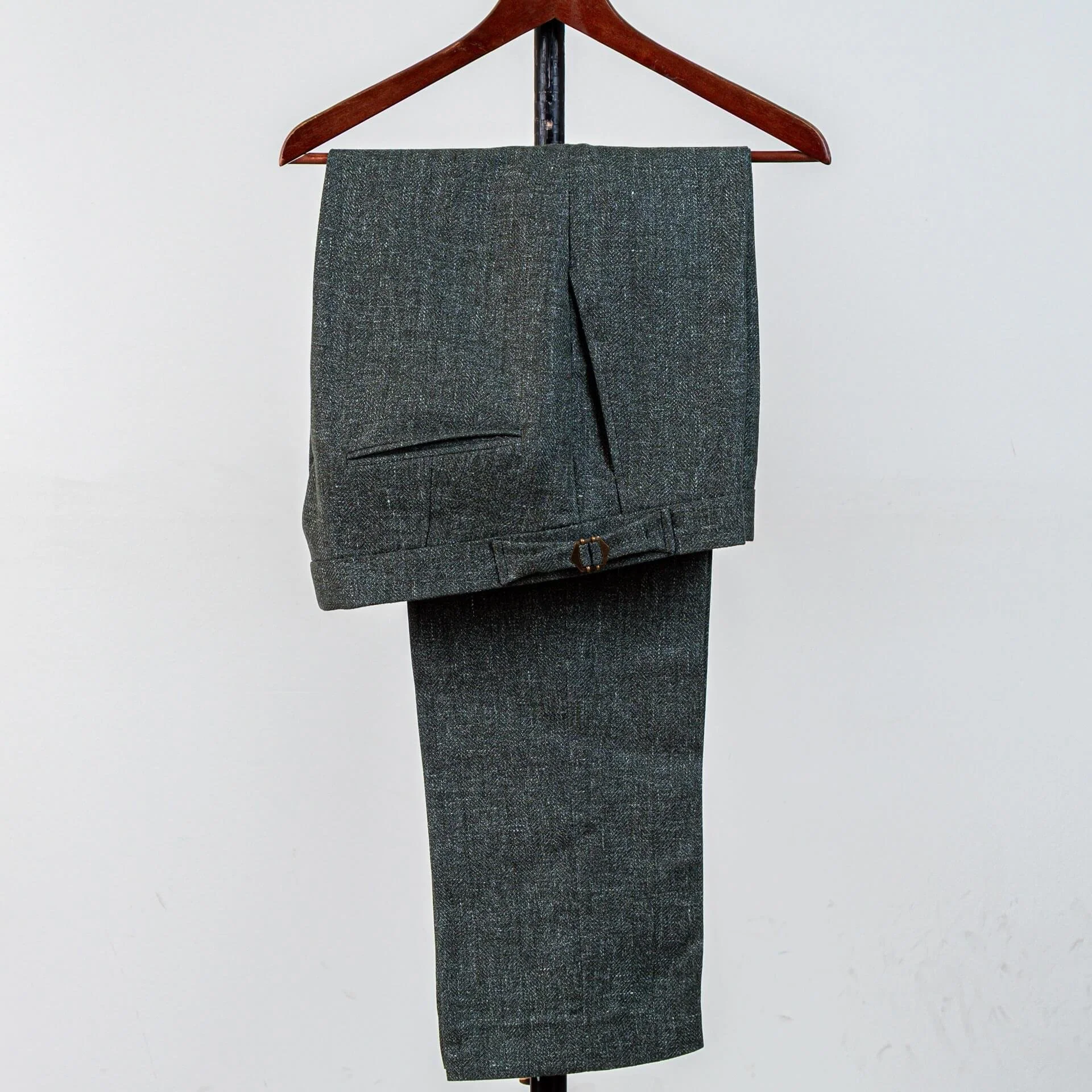 Gevouwen groene trouwpantalon in wol-linnen-zijde op houten hanger. Zichtbaar: steekzakken, bronzen riemgesp en natuurlijke textuur. Licht en ademend voor zomerbruiloft.