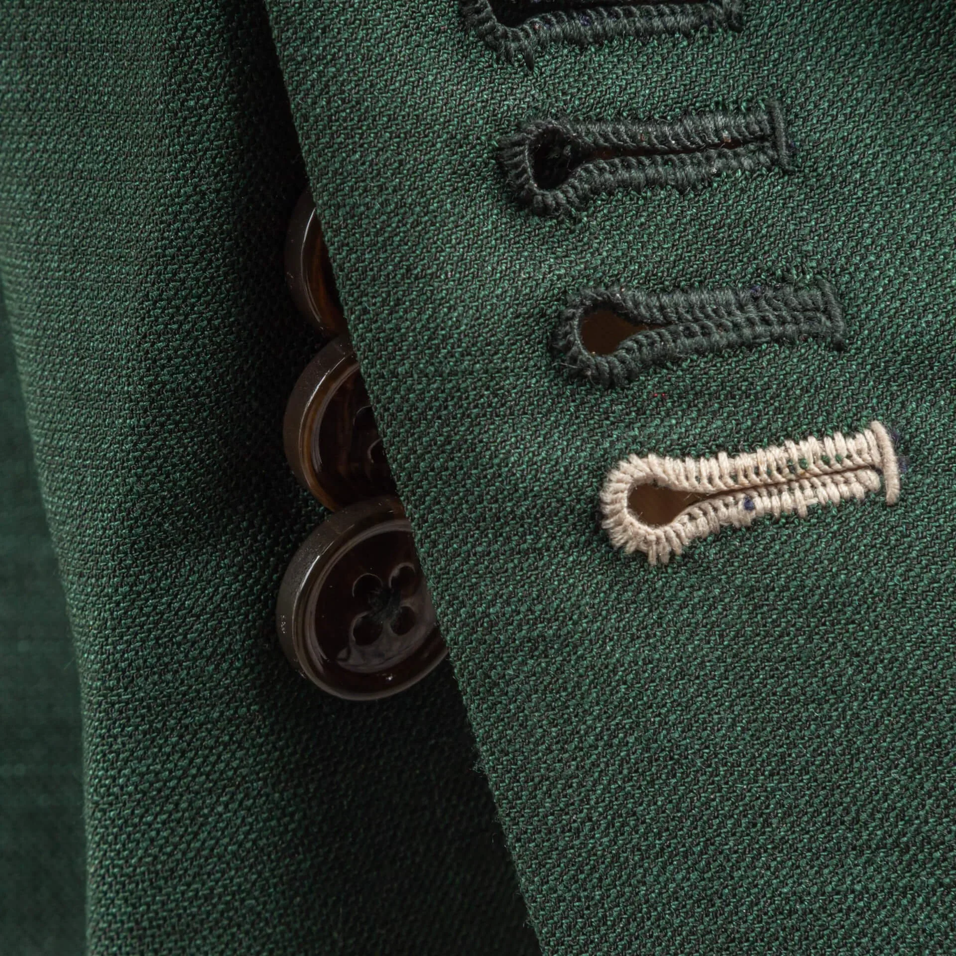 Detail van mouwknopen op saffiergroen trouwpak met werkende knoopsgaten, onderste knoopsgat gestikt in contrasterende zandkleur als persoonlijk accent, donkerbruine hoornen knopen