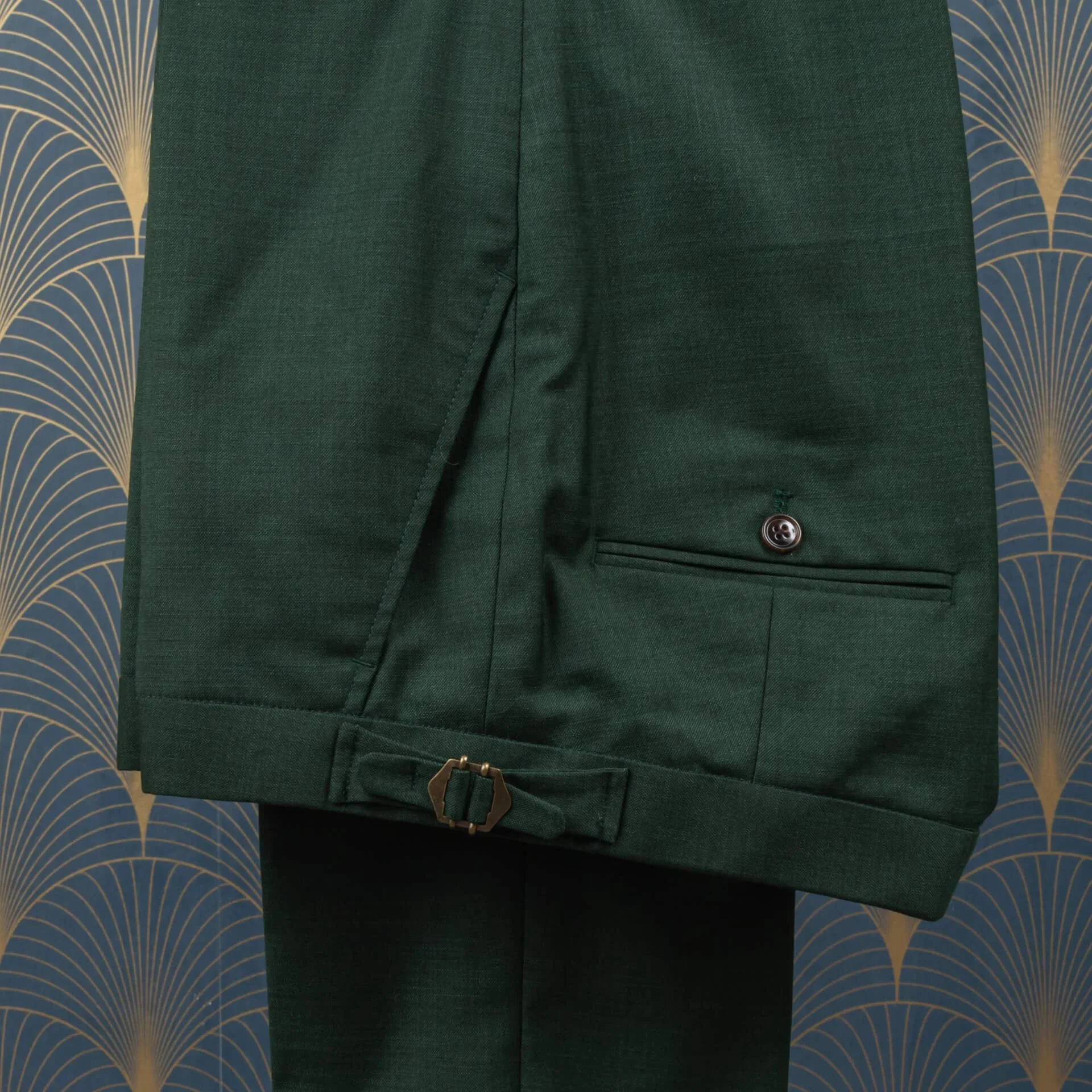 Close-up van saffiergroene merino wol pantalon toont klepzak met donkerbruine knoop en koperen side adjuster, 280 gram sharkskin stof