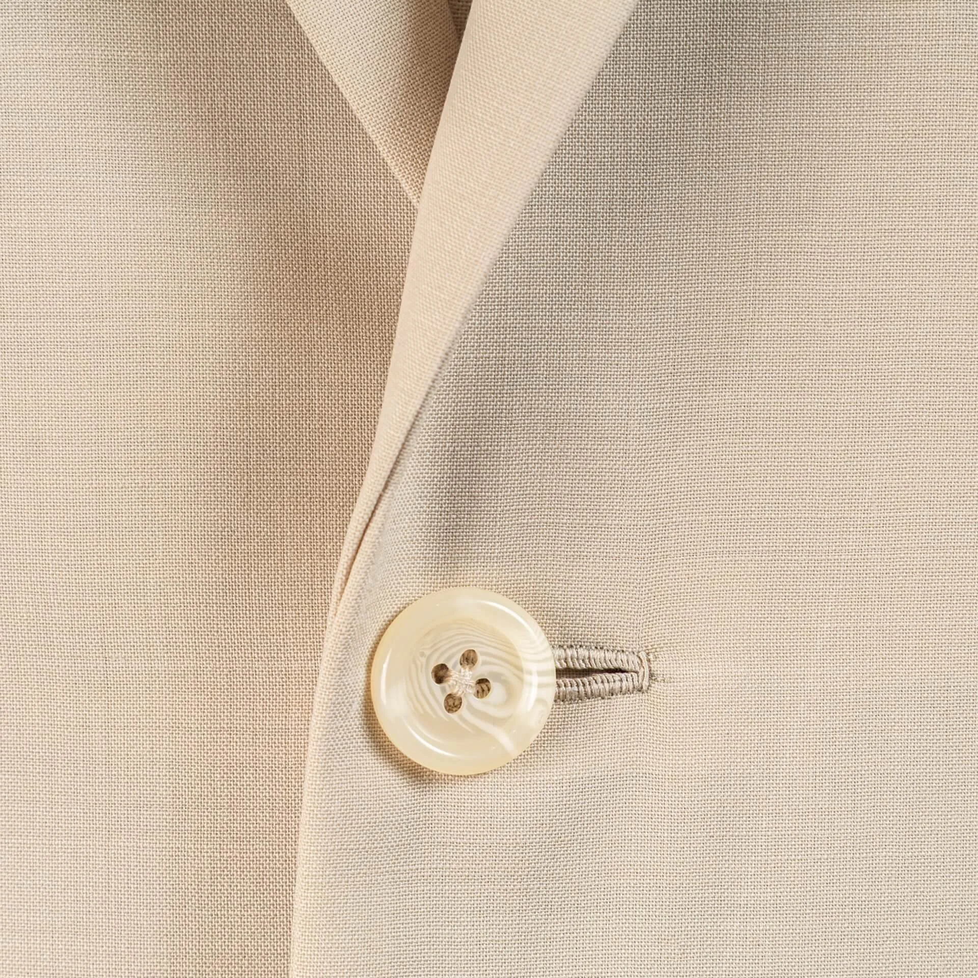 Detail van crème hoornen knoop met handgestikt knoopsgat op biscuit trouwpak, plain weave woltextuur en vakmanschap zichtbaar