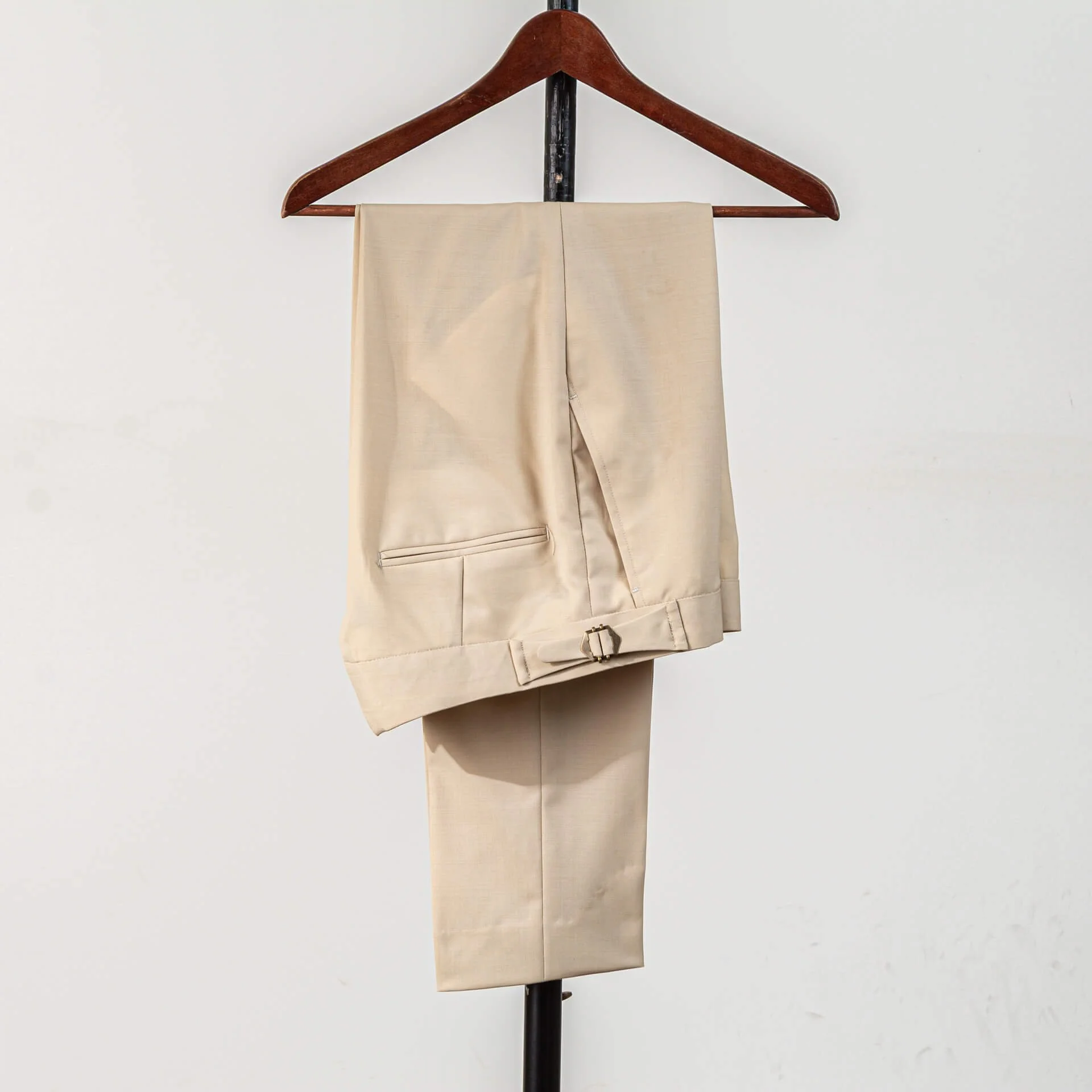 Pantalon op maat in biscuit kleurige wol van Bateman & Ogden met side adjusters en messing gesp voor verstelbare pasvorm, paspelzak zichtbaar, op houten kledinghanger