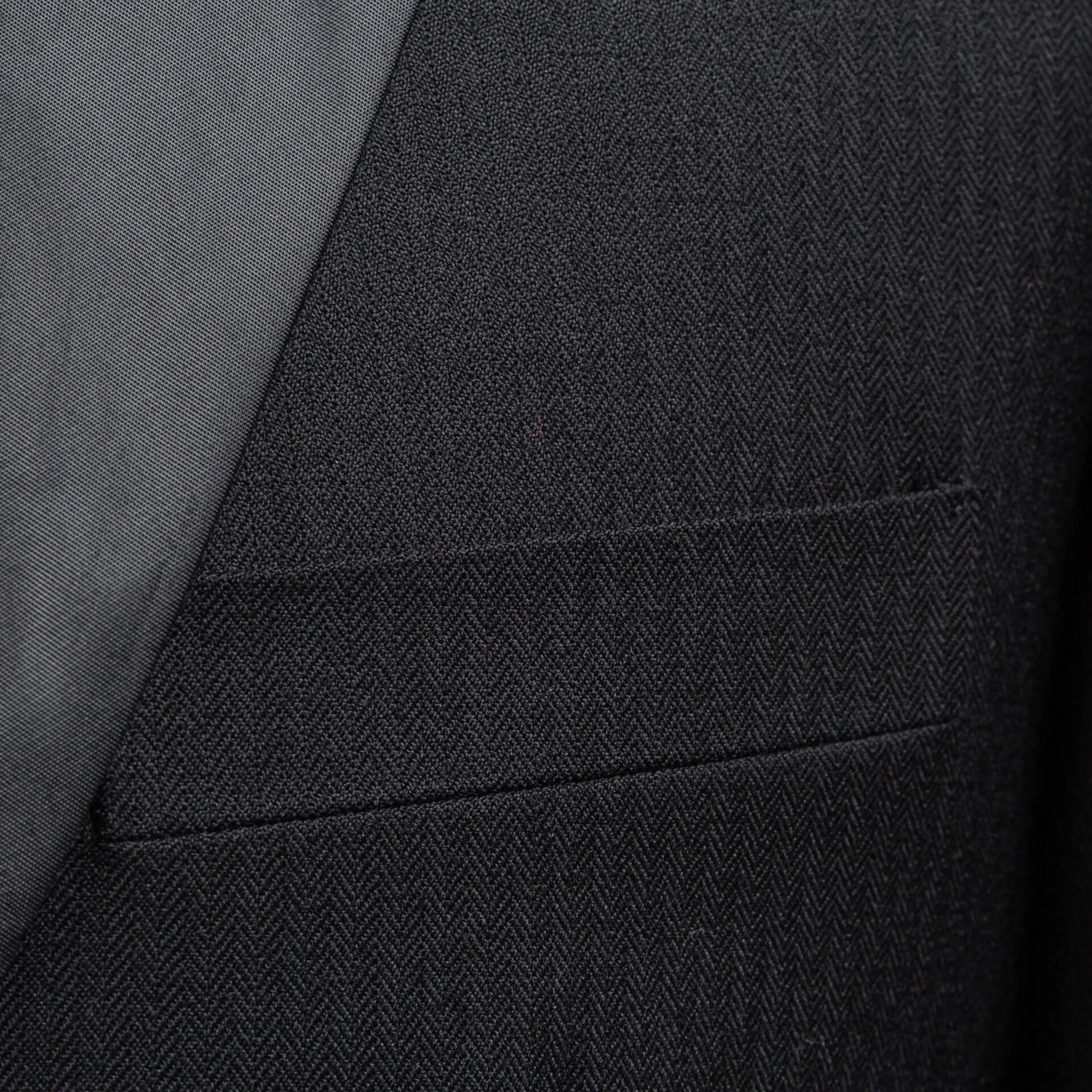 Close-up van de peak revers met satijnen afwerking en borstzak van een smoking voor het trouwen op maat in zwarte visgraat, Super 100's wol en cashmere Holland & Sherry Cape Horn