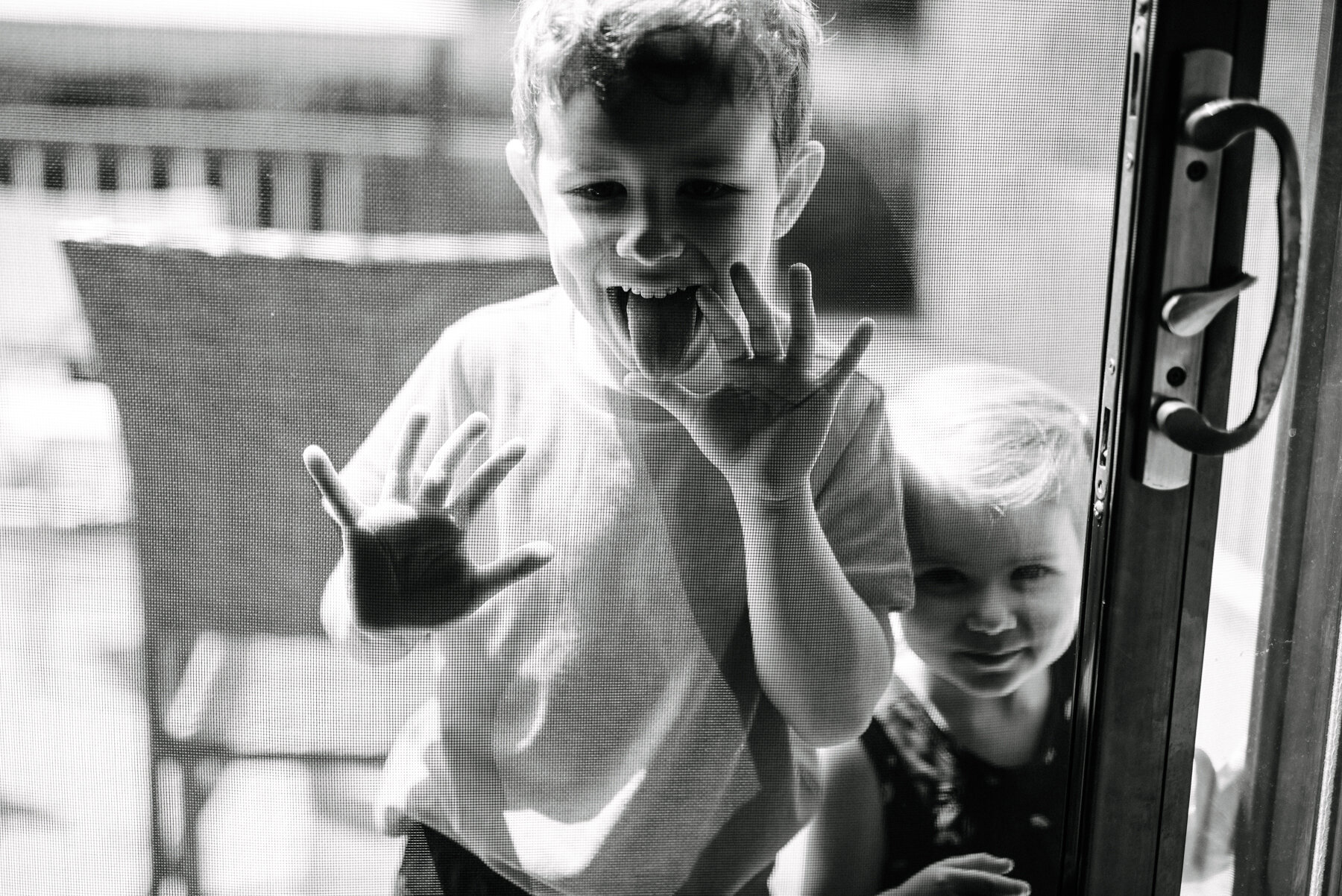 026photo-noir-et-blanc-enfants-qui-grimacent-a-travers-moustiquaire-porte-patio-photographe-de-famille-a-montreal-marianne-charland-0781.jpg