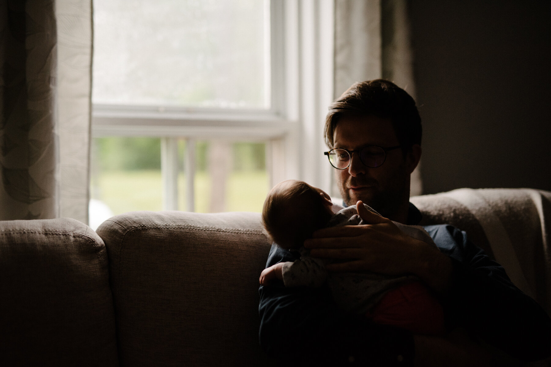 026photo-papa-qui-tient-et-regarde-tendrement-bebe-assis-sur-divan-devant-une-fenetre-photographe-de-nouveau-ne-a-montreal-marianne-charland-231.jpg