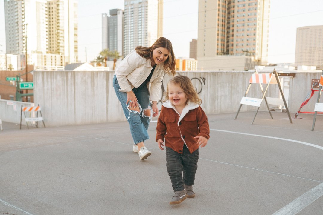 A woman and a young girl, outdoors on a rooftop parking lot with city buildings in the background, smiling and walking.
