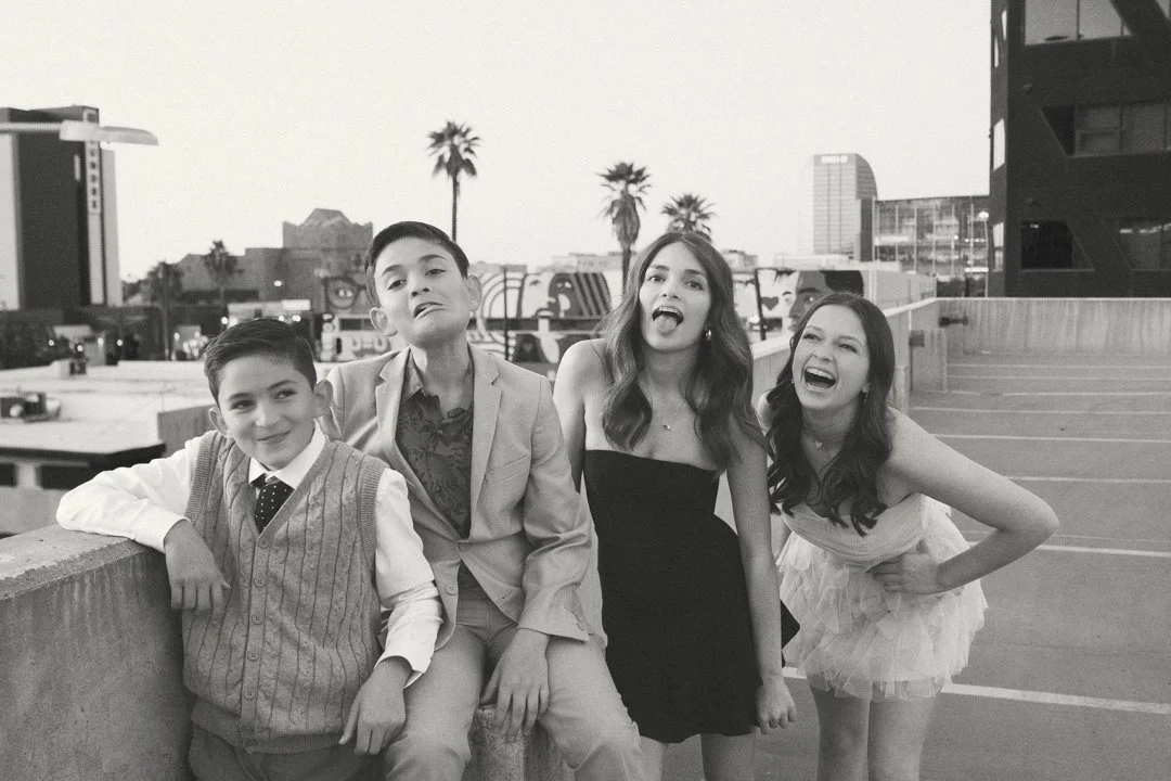 Four children standing on a rooftop parking lot, with city buildings and palm trees in the background, dressed in semi-formal attire.